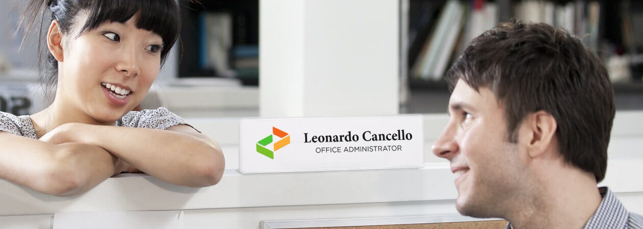 A man and woman make eye contact in an office, with a plastic name plate visible.
